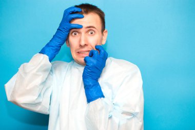 Koruyucu giysili korkmuş bir adamın portresi, mavi arka planda virüslere karşı tıbbi maske ve eldivenler ve enfeksiyonlara karşı el ele kapalı..