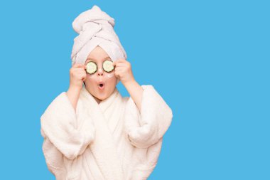 Beyaz önlüklü, maskeli, deri yüzlü ve mavi arka planda gözlerinde salatalık olan komik küçük kız. Spa cilt bakımı