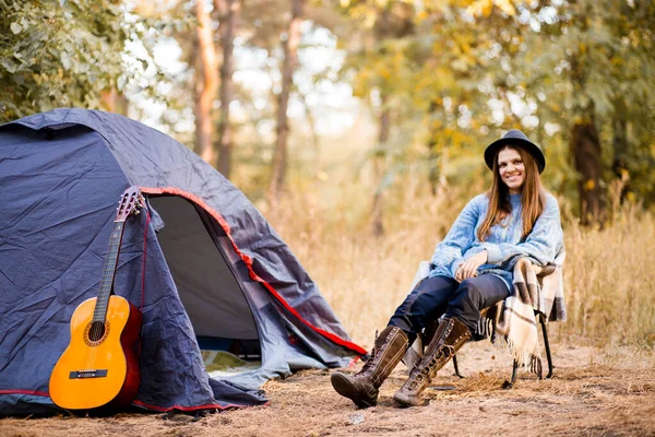 Young woman in warm sweater and black hat resting with guitar near camping tent in wilderness autumn forest