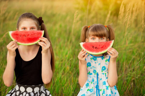 Two funny little sisters eating and hide face by watermelon outdoors on warm and sunny summer day. Healthy organic food for little kids.