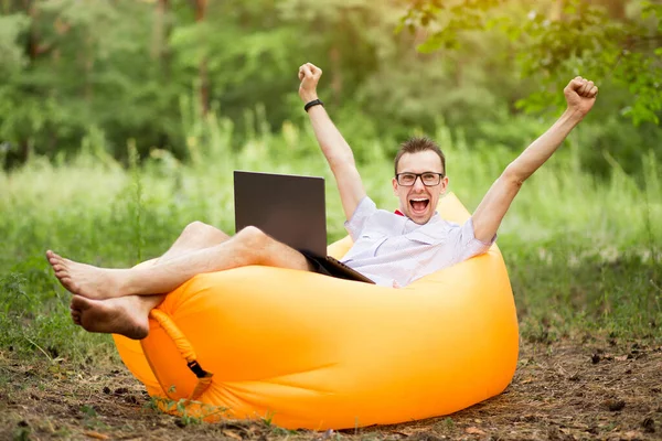 Barefoot man with hands up in winner pose and happy screaming, in shirt, red bow tie and glasses lying on inflatable sofa with laptop in summer park.