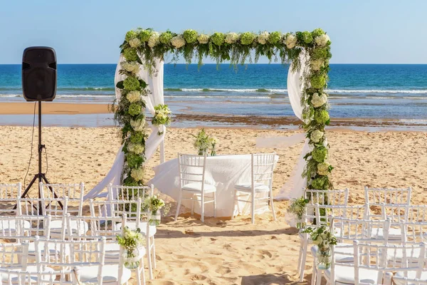 Romantyczna ceremonia ślubna nad oceanem, plaża w słoneczny dzień. Drewniany łuk ozdobiony świeżymi kwiatami. Białe fotele na piasku. — Zdjęcie stockowe