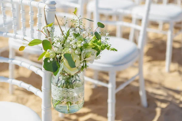 Vita stolar vid en bröllopsceremoni på stranden, dekorerade med en hängande burk med blommor, en solig dag. Närbild. — Stockfoto