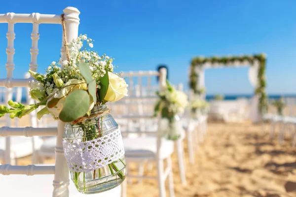Luxuriöse Hochzeitszeremonie am Meer, am Strand. Weiße Stühle dekoriert mit einem schönen Blumenstrauß in einem Gefäß, das an ihnen hängt. — Stockfoto
