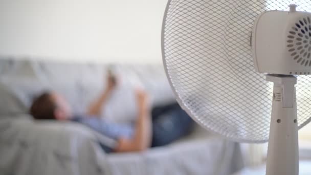 Elektrischer Ventilator im Raum zur Luftkühlung. Ein Mann liegt mit einem Handy in der Hand auf einem Sofa. Nahaufnahme. — Stockvideo