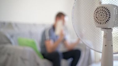 Kanepede oturan bir adam, telefona bakıyor, akıllı telefon. Isıyı düşürmek için vantilatör de dahil. Bulanık arkaplan.