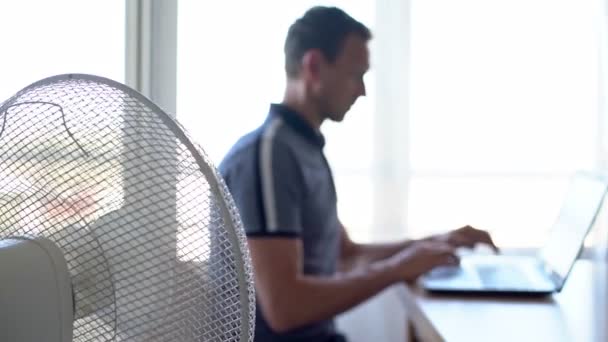 Um homem em um computador, laptop em um ambiente de trabalho, com o ventilador elétrico ligado. Fundo desfocado . — Vídeo de Stock