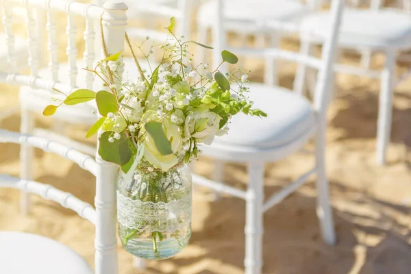 Sedie bianche a una cerimonia di nozze in spiaggia, decorate con un barattolo appeso di fiori, in una giornata di sole . — Foto Stock