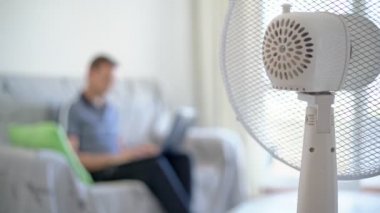 Bilgisayarı olan bir adam, ev ortamında çalışan bir dizüstü bilgisayar, kanepede oturuyor, tişörtünü çıkarıyor. Elektrikli vantilatör dahil, ısıdan korunuyor. Bulanık arkaplan.