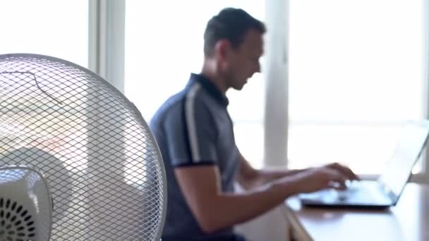 Ein Mann am Computer, Laptop in der häuslichen Arbeitsumgebung, den elektrischen Ventilator eingeschaltet, um Wärme zu sparen. Unklarer Hintergrund. — Stockvideo