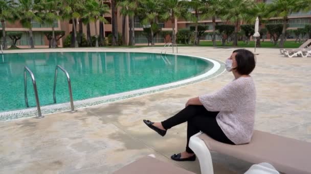 Coronavirüs maskeli, alerjisi olan orta yaşlı bir kadın otel havuzunun yanındaki güneşli bir yatakta oturuyor. Yakın plan.. — Stok video