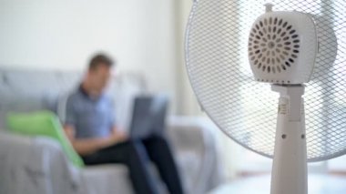Bilgisayarı olan bir adam, ev ortamında çalışan bir dizüstü bilgisayar, kanepede oturuyor. Elektrikli vantilatör dahil, ısıdan korunuyor. Bulanık arkaplan.