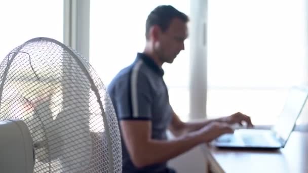 Un uomo al computer, un portatile in un ambiente domestico di lavoro, con la ventola elettrica accesa, risparmiando calore. Sfondo sfocato . — Video Stock