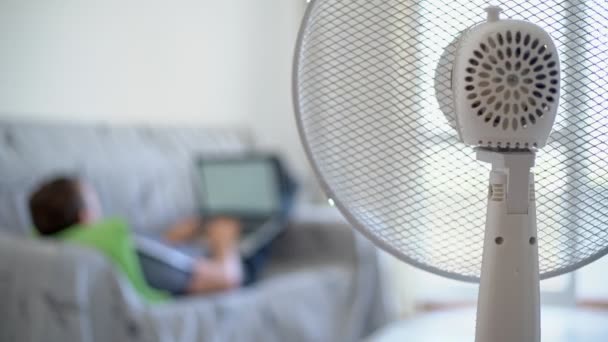 Egy férfi fekszik otthon egy kanapén egy számítógéppel. Bekapcsolt az elektromos ventilátor. Homályos háttér. — Stock videók