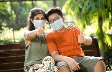 Mother and son with protective face mask giving thumbs up at park clipart