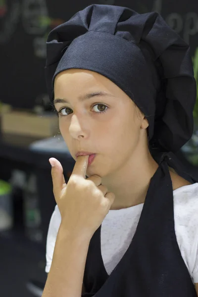 Beautiful young cook chef girl in a black chef\'s cap licks finger