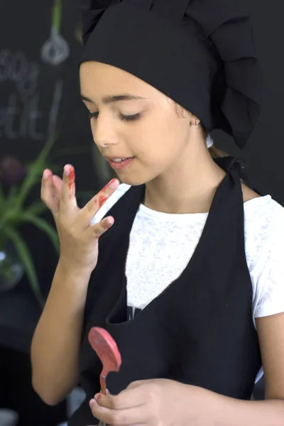 Beautiful young cook chef girl in a black chef's cap licks finger