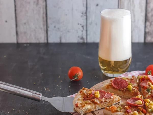 Pizza with corn, sausage, tomatoes on wooden board and light beer with foam in a glass