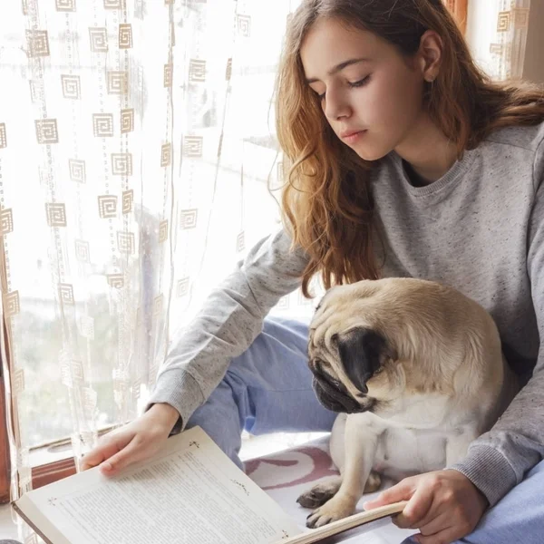 Ładna dziewczyna z jej pies mops, czytanie książki w pobliżu okna — Zdjęcie stockowe