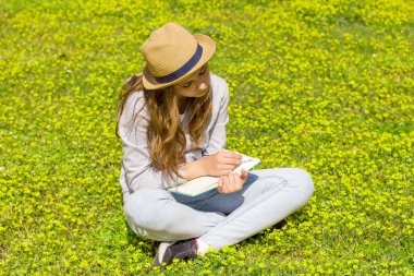 Sevimli Genç kız yeşil bir çayır üzerinde bir kağıt pad üzerinde notlar yazıyor. Seçici odak