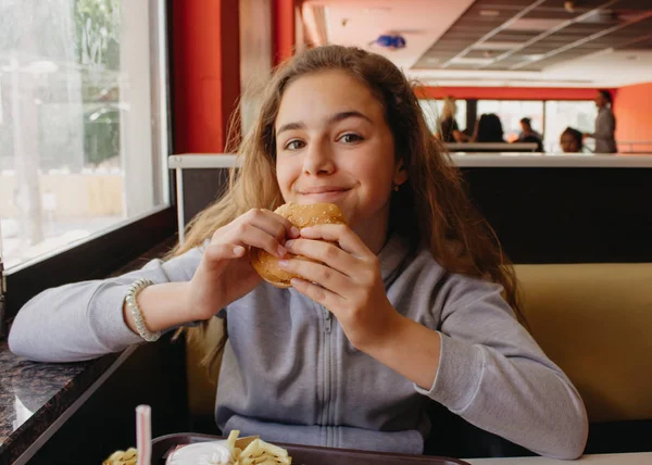 Csinos fiatal tini lány étvágya eszik egy hamburger egy kávézóban — Stock Fotó