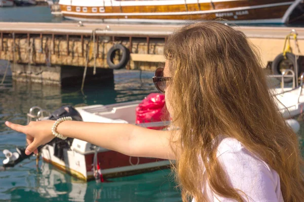 Young girl with long hair sits on a pier in the yacht port and shows her hand on the blue sea — Stockfoto