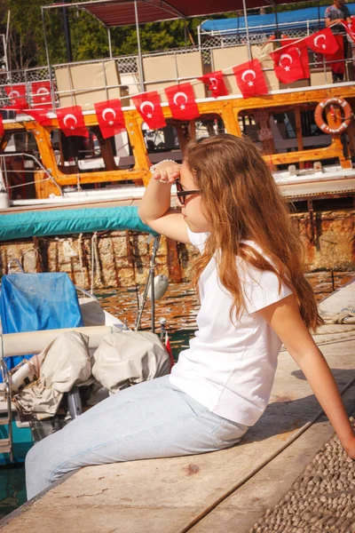 Mladá dívka s dlouhými vlasy sedí na molu v přístavu jachty a dívá se na modré moře — Stock fotografie