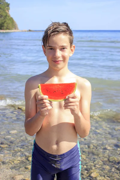 Söt tonåring pojke innehar en bit vattenmelon på stranden vid havet — Stockfoto