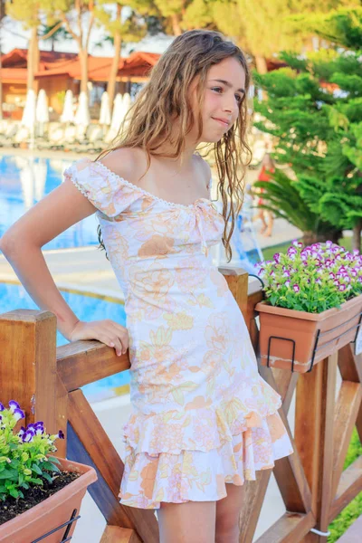 Linda chica adolescente en un vestido romántico y con el pelo largo está de pie en el puente junto a la piscina —  Fotos de Stock