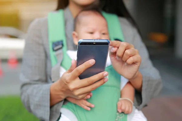 Close-up business mother in suit is using smartphone with carrying her baby boy in hipseat.