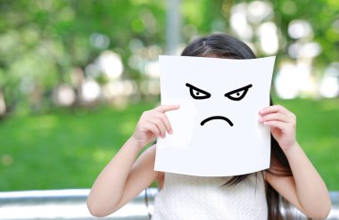Little child girl holding up a white paper with draw emotion angry face in nature park. clipart