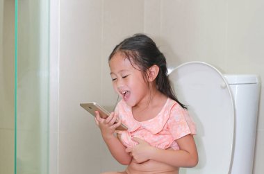 Tuvalette otururken ve smartphone oynamaya yakın çekim mutlu küçük Asyalı çocuk kız.