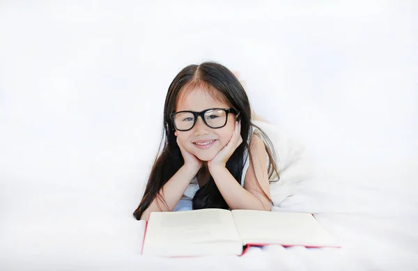 アジア子供女の子眼鏡を白い背景に ベッドに横になっているハードカバーの本を読んで — ストック写真