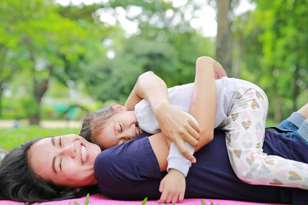 快乐的亚洲妈妈拥抱躺在绿色草坪上的女儿 有趣的母亲和女孩在夏季公园玩耍 — 图库照片