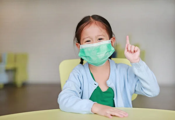 Cute little Asian child girl wearing a protective mask with showing one forefinger sitting on kid chair in children room.