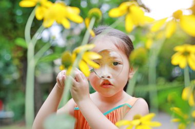 Pretty little Asian child girl with magnifying glass looks at flower in summer park. clipart