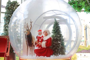 Mutlu küçük kız çocuk ve annesi olarak Noel Baba kostüm Giydir vardır eğlenceli büyük kış kar küresi Ren geyiği ile Noel zamanında. Neşeli Noel ve mutlu yeni yıl.