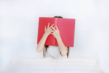 Küçük Asyalı çocuk kız yatakta yastık ile oturan açık ciltli kitap ile yüz gizleme.
