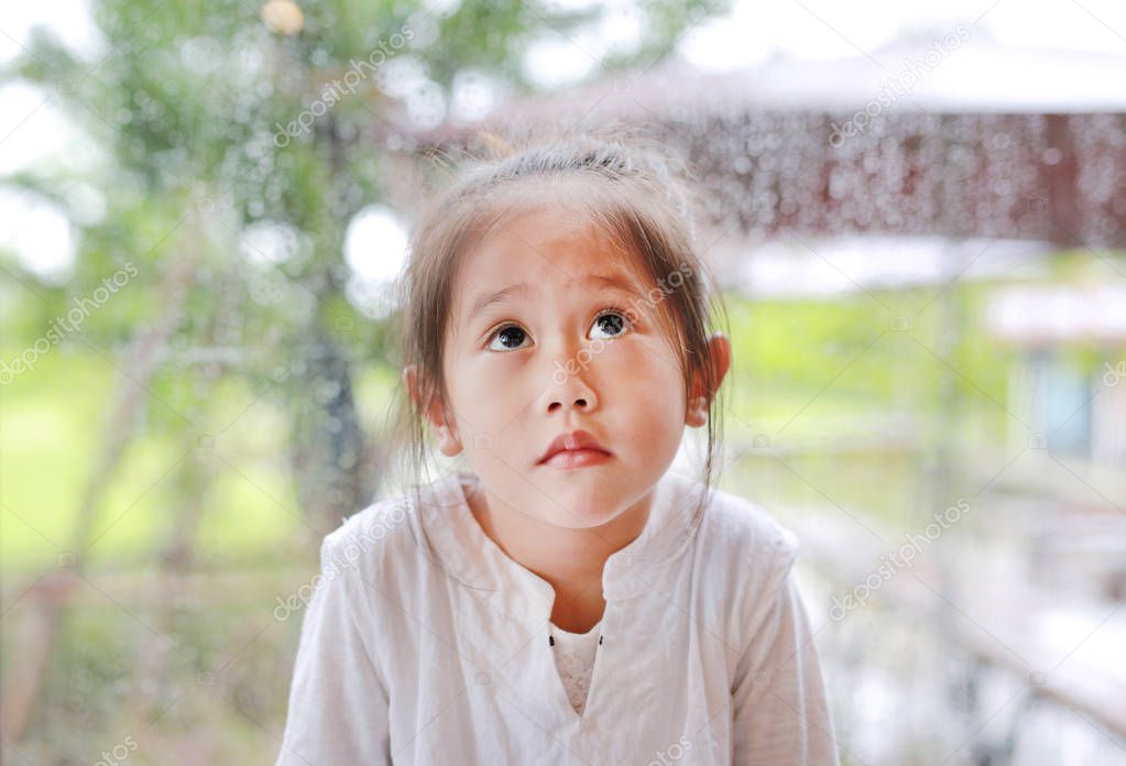 Little Asian child girl gesture with peaceful face and looking up against glass window with water drop at rainy day.