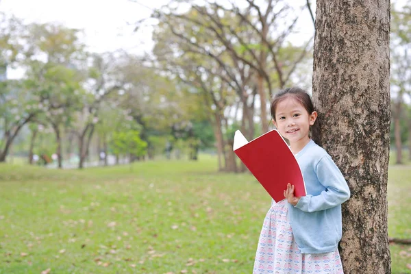 肖像画アジア子供女の子立ってカメラを探して木の幹に対して無駄のない屋外の公園で本を読んで — ストック写真