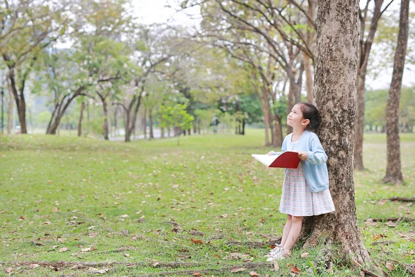 小さな子供の女の子立って空を見ながら木の幹に対して無駄のない屋外の夏の公園で本を読んで — ストック写真