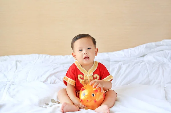 家でベッドに座っているピギーバンクと伝統的な中国のドレスを着た小さなアジアの男の子 子供貯蓄お金の概念 — ストック写真