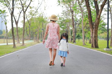 Anne ve kızı yolda yürürken ve açık doğa bahçesinde el ele tutuşuyor. Geri görünüm.