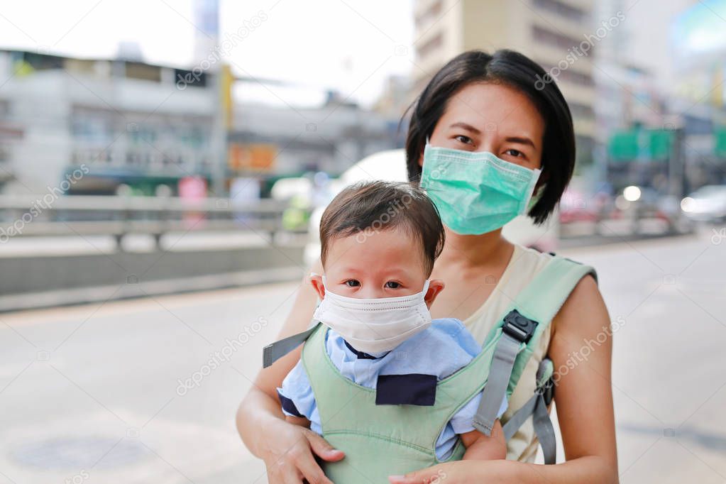 Asian mom carrying her baby by hipseat walking with wearing a protection mask against air pollution in Bangkok city. Thailand.