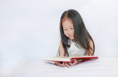 Gülümseyen küçük Asyalı çocuk kız yastık yatak beyaz arka plan üzerinde yatan ciltli kitap okuma.