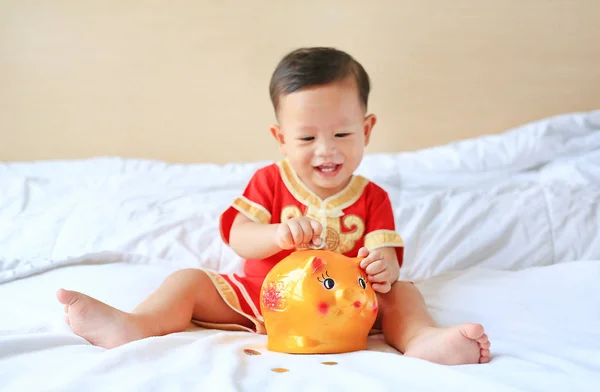 面带微笑的亚洲小男孩身着中国传统服装 把一些硬币放进一个坐在家里床上的小猪银行里 孩子省钱的概念 关注小猪银行 — 图库照片