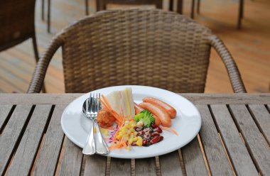 Ahşap masada beyaz bir tabakta sandviç, sosis ve salata ile kahvaltı.