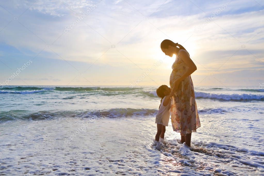 Silhouette daughter embracing pregnant mother relaxing on the beach at sunset.