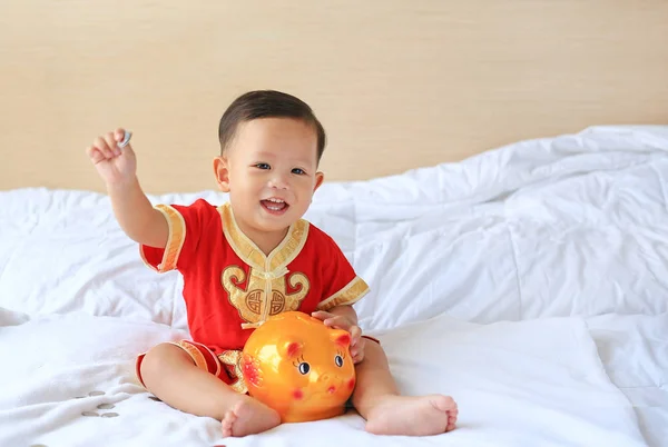 面带微笑的亚洲小男孩身着中国传统服装 把一些硬币放进一个坐在家里床上的小猪银行里 孩子省钱的概念 — 图库照片