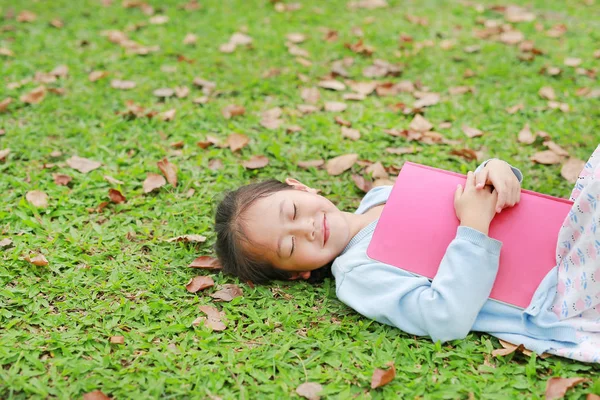 夏の庭で乾燥した葉と緑の草の上に横たわっている本を持つ幸せな小さな女の子の女の子 — ストック写真
